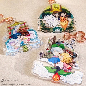 May include: Three colorful acrylic charms featuring characters from popular anime films. The first charm features a white and blue dragon with two girls on its back. The second charm features a white wolf and a red deer with two girls in the background. The third charm features a flying castle with two girls in the foreground.