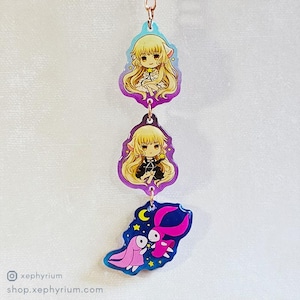 Puede incluir: Un charm de tres piezas con ilustraciones de estilo anime. Los dos charms superiores representan a un personaje rubio con diferentes atuendos, mientras que el charm inferior muestra una criatura rosa y roja sobre un fondo azul. Los charms están conectados por una cadena de color cobre.