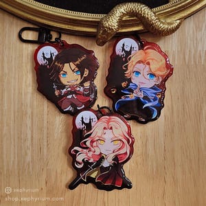 May include: Three anime-style character keyrings with red borders and black accents. Each keyring features a different character with blue eyes, set against a backdrop of a full moon and a silhouette of a castle. The keyrings are on a wooden surface.