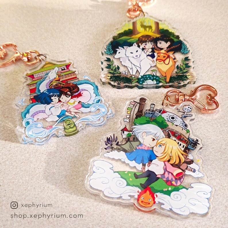 May include: Three colorful acrylic charms featuring characters from popular anime films. The first charm features a white and blue dragon with two girls on its back. The second charm features a white wolf and a red deer with two girls in the background. The third charm features a flying castle with two girls in the foreground.