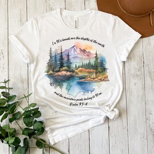 May include: White t-shirt with a watercolor illustration of a mountain range, lake, and trees. The text "In His hands are the depths of the earth and the mountain peaks belong to Him. Psalm 95:4" is printed on the shirt.