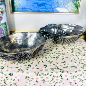 May include: A silver-toned, two-part serving dish with a snail-shaped handle. The dish has a textured, bark-like exterior and a smooth interior. The snail handle is detailed with ridges and a shell. The dish sits on a floral patterned surface.
