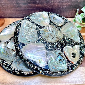 May include: Two round black coasters with iridescent mother of pearl shell pieces embedded in the resin. The coasters are stacked on top of each other.