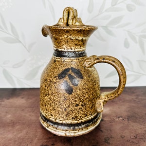 May include: A brown ceramic pitcher with a speckled glaze and a black and white design. The pitcher has a handle and a lid.