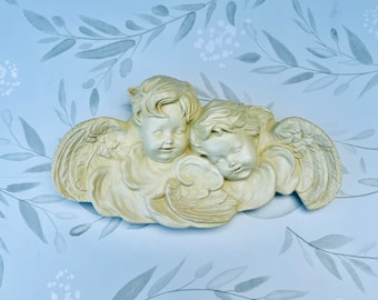 May include: A white plaster wall decor featuring two cherubs with wings, sleeping on a cloud. The cherubs are sculpted in a realistic style with delicate features.