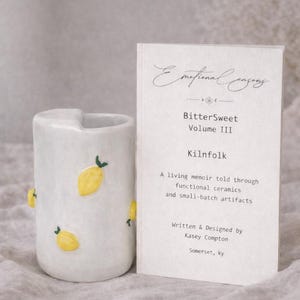 Puede incluir: Una taza de cerámica blanca con un asa negra y el texto "But First, Coffee" en un estilo manuscrito. La taza está sobre una superficie blanca. La taza mide aproximadamente 10 cm de alto.