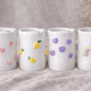 Puede incluir: Cuatro porta cepillos de dientes de cerámica blanca con diseños decorativos. Uno tiene corazones rosas, otro limones amarillos, un tercero flores moradas y el último un arcoíris. Cada soporte tiene una pequeña abertura en la parte superior.