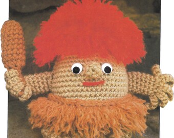 May include: A crocheted toy with red hair, brown fur, and large white eyes. The toy is holding a brown stick in its right hand.