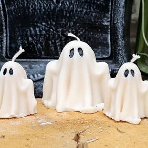May include: Three white ghost-shaped candles with black oval eyes and wicks. The candles vary in size, with the largest in the centre. The candles are on a wooden surface, with a dark background and a green lantern visible.