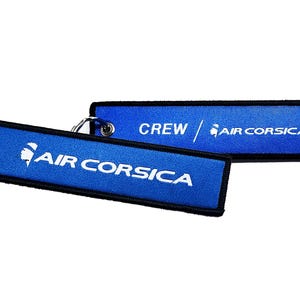 Peut inclure: Porte-clés en tissu bleu et noir avec un logo blanc qui dit "AIR CORSICA" et "CREW / AIR CORSICA".