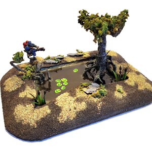 May include: A miniature tabletop gaming terrain featuring a small pond with green lily pads, a miniature figure, and a small tree with brown roots. The pond is surrounded by a brown dirt base with a sandy texture.