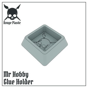 以下が含まれることがあります： 骸骨と交差した骨のデザインが施された灰色のプラスチック製の接着剤ホルダー。ホルダーは、縁が盛り上がった正方形の形をしています。ホルダーの下には「Mr Hobby Glue Holder」という文字が印刷されています。