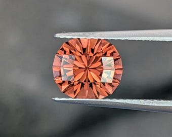May include: A faceted, round gemstone held by tweezers. The gem is a warm orange color with intricate internal facets that reflect light. The tweezers are silver-colored, and the background is a dark gray.