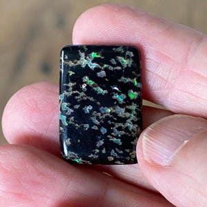 May include: A rectangular black gemstone with a polished surface featuring scattered, irregular inclusions of iridescent green and blue flecks that create a vibrant, speckled pattern across the dark, opaque base of the stone.