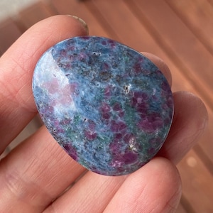 May include: A polished, oval-shaped gemstone with a mix of colors. The stone features a base of blue and green hues, speckled with vibrant red spots. The smooth surface of the stone reflects light, highlighting its natural patterns.