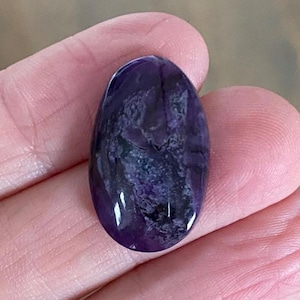 May include: A polished, oval-shaped gemstone with a deep purple hue, featuring swirling patterns of lighter purple and dark gray. The stone is smooth and reflective, showcasing natural variations in color and texture.
