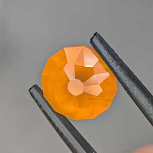 以下が含まれることがあります： 黒い金属製のピンセットで保持された、円形でファセットカットされたオレンジ色の宝石。石の中央には円形のファセットがあり、その周囲を複数の三角形や台形のファセットが囲むことで、表面に幾何学的な星のようなパターンを作り出しています。
