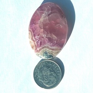 May include: A polished, oval-shaped gemstone with pink and white marbling. The stone is next to a U.S. dime coin for size comparison. The gemstone has a smooth, glossy surface and is set against a white background.