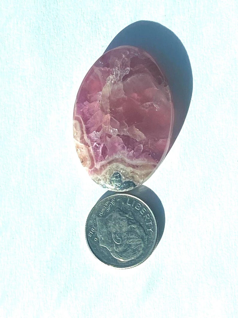 May include: A polished, oval-shaped gemstone with pink and white marbling. The stone is next to a U.S. dime coin for size comparison. The gemstone has a smooth, glossy surface and is set against a white background.