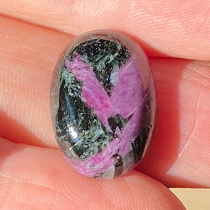 May include: An oval-shaped cabochon gemstone featuring a polished surface with a distinct pattern of vibrant magenta-pink inclusions set against a dark, mottled forest green and black background, showcasing natural mineral variations.