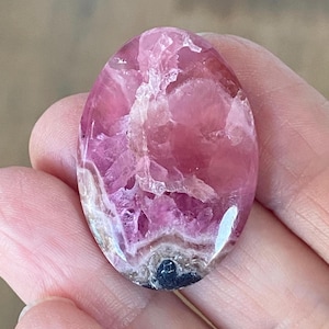 May include: Oval-shaped pink gemstone with a polished surface. The stone has a mix of light and dark pink hues, with some areas appearing translucent. The gemstone is held in a hand, showcasing its size and shape.