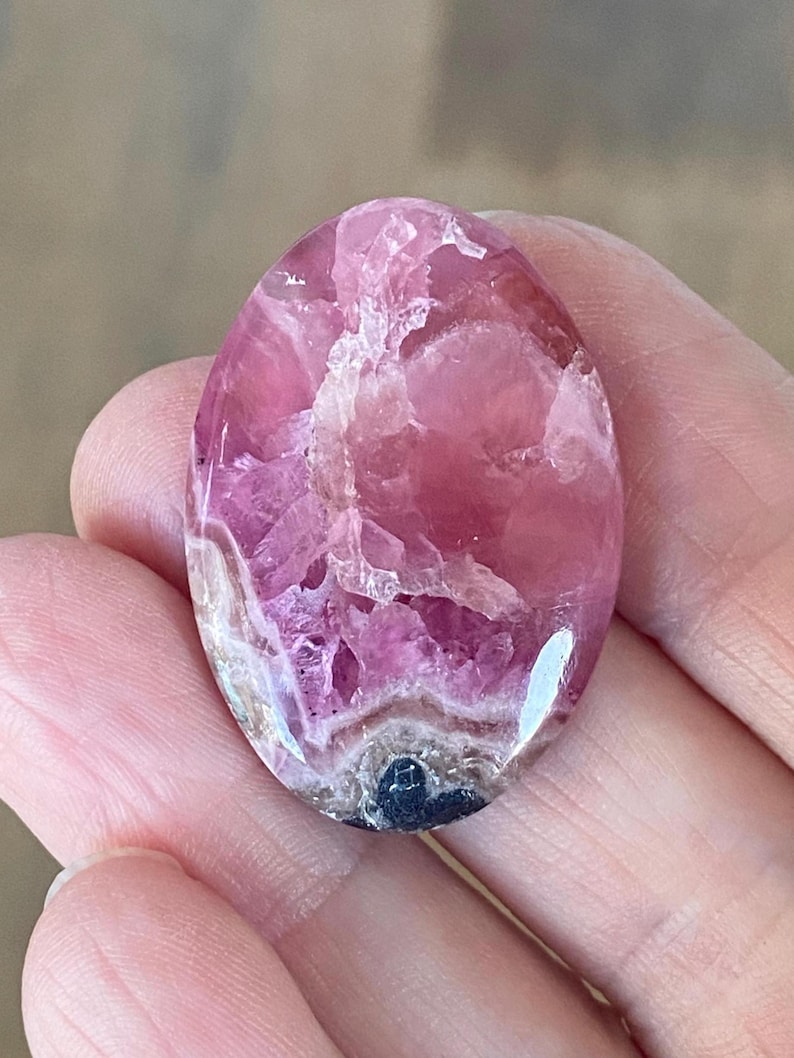May include: Oval-shaped pink gemstone with a polished surface. The stone has a mix of light and dark pink hues, with some areas appearing translucent. The gemstone is held in a hand, showcasing its size and shape.