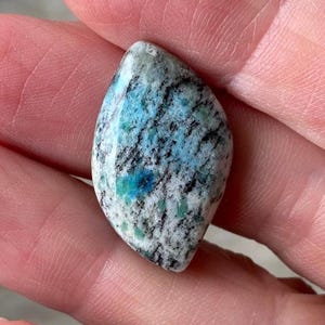 May include: A polished, marquise-shaped gemstone featuring a mottled pattern of white, grey, and black mineral inclusions interspersed with vibrant patches of bright turquoise and deep blue color throughout the smooth, rounded surface.