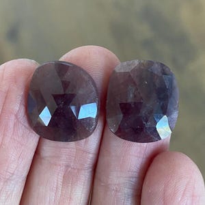 May include: Two faceted, dark purple gemstones with rounded edges are held in a hand. The gems have a polished, reflective surface, catching the light and displaying a range of dark purple hues. The gemstones are likely for jewelry making.