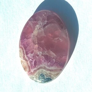 May include: Oval-shaped polished gemstone with a mix of pink and white hues. The stone has a translucent quality, with visible crystalline structures and a smooth, glossy surface. The gemstone is set against a white background.