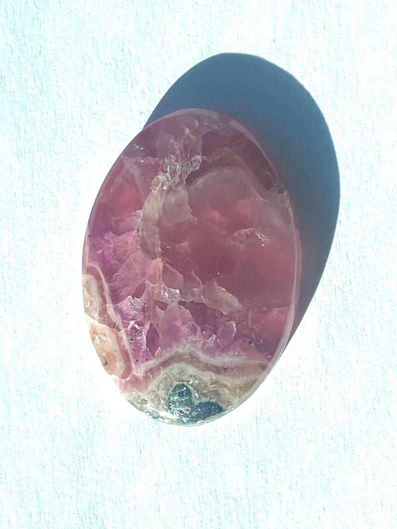 May include: Oval-shaped polished gemstone with a mix of pink and white hues. The stone has a translucent quality, with visible crystalline structures and a smooth, glossy surface. The gemstone is set against a white background.