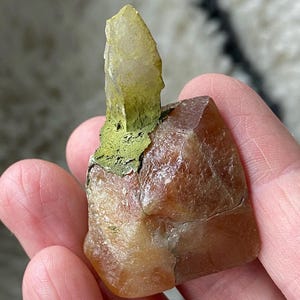 May include: A natural crystal formation featuring a large, faceted, amber-colored crystal base with a smaller, light green crystal protruding from the top. The crystals are held in a hand, showcasing their natural beauty.