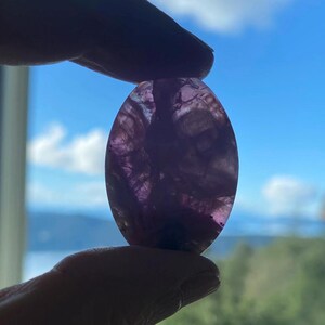 May include: An oval-shaped, translucent purple gemstone is held up against a bright blue sky with fluffy white clouds. The stone has a marbled appearance with varying shades of purple and white. The gemstone is being held between two fingers.
