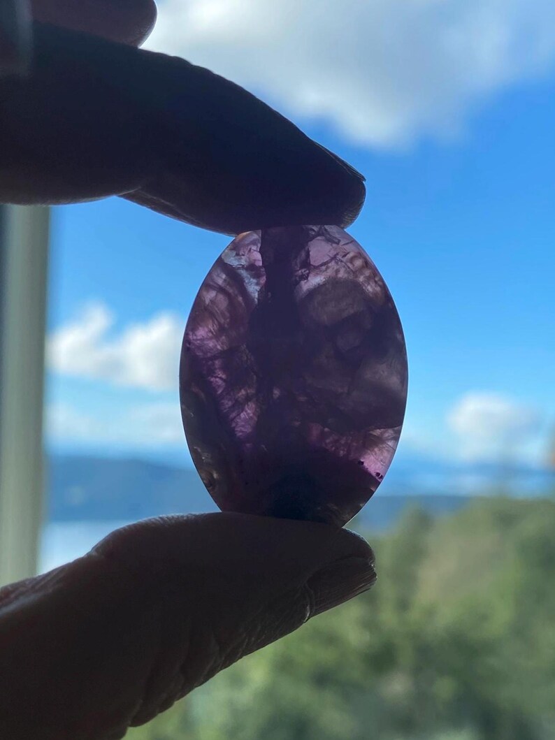 May include: An oval-shaped, translucent purple gemstone is held up against a bright blue sky with fluffy white clouds. The stone has a marbled appearance with varying shades of purple and white. The gemstone is being held between two fingers.