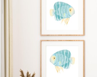 Könnte beinhalten: Zwei gerahmte Aquarelldrucke eines tropischen Fisches. Der Fisch ist hellblau mit weißen Streifen und gelben Akzenten. Die Rahmen sind aus hellbraunem Holz, und der Hintergrund ist weiß.