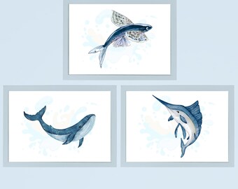 Könnte beinhalten: Drei gerahmte Aquarelldrucke mit Meeresmotiven. Der obere Druck zeigt einen fliegenden Fisch mit detaillierten Flügelmustern. Der untere linke Druck zeigt einen Wal, und der untere rechte Druck zeigt einen Marlin. Alle sind in Blautönen gehalten.