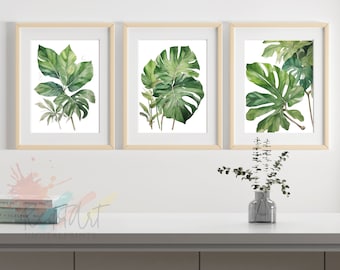 May include: Three framed watercolor paintings of green tropical leaves on a white wall. The paintings are in wooden frames and are arranged in a row. There is a small vase with green leaves on a white countertop below the paintings.