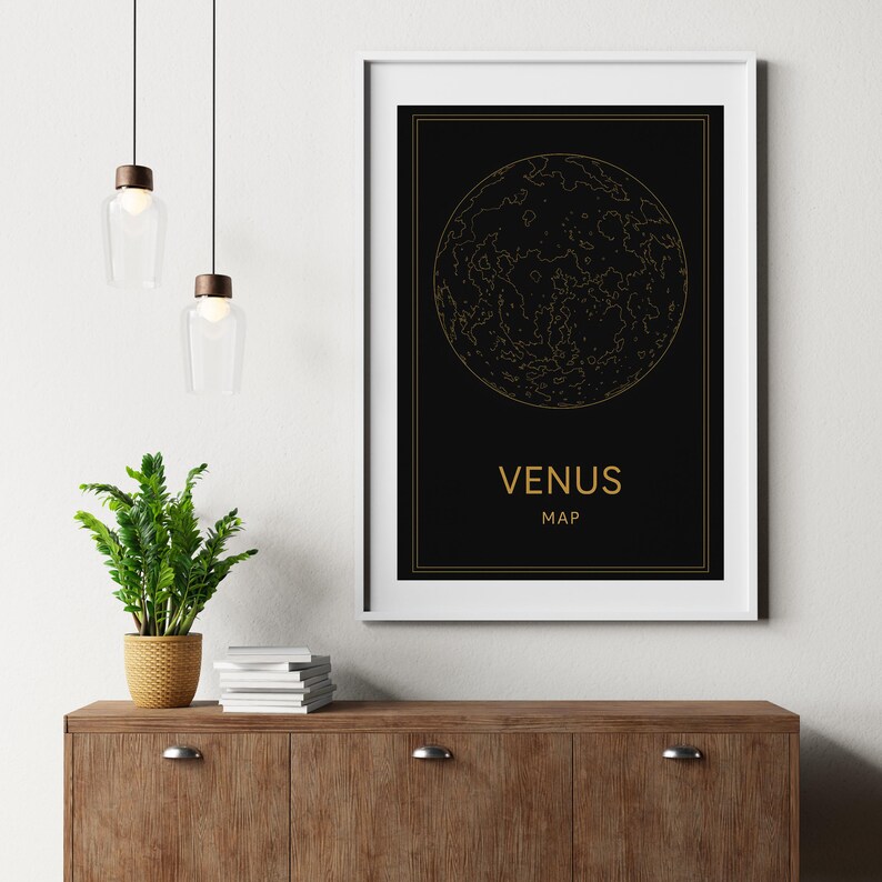 以下が含まれることがあります： 金色の文字で「VENUS MAP」と書かれた、金星の地図の白黒プリント。プリントは白いフレームで額装され、白い壁にかけられています。