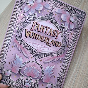 Może przedstawiać: Prostokątny przedmiot w kolorze lawendy z napisem "FANTASY WONDERLAND" w złotych literach. Projekt zawiera motywy kwiatowe i gwiazd, z dekoracyjną obwódką. Przedmiot jest trzymany w dłoni.