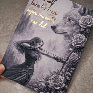 May include: A monochrome illustration depicts a woman with a bow and arrow, a wolf, and roses. Gold text reads "The wolf knew her heart before the world ever did." The artwork has a fantasy theme.