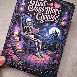 Puede incluir: Impresión rectangular con un esqueleto leyendo un libro, con el texto "Just One More Chapter". La imagen muestra un gato negro, un castillo, fantasmas, rosas y una luna llena sobre un cielo estrellado.