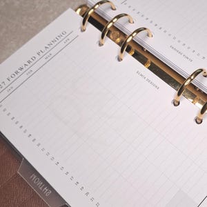 Op de afbeelding: Bruin leren planner met gouden ringen en een pen. De planner is open op een pagina met rasterlijnen en de tekst "2027 Forward Planning". De planner is ontworpen voor organisatie en planning.
