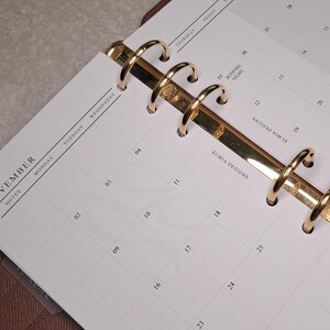 May include: A brown leather planner open to a November 2026 calendar page. The planner features gold-coloured rings and a pen holder. The calendar pages are white with black text and numbers.