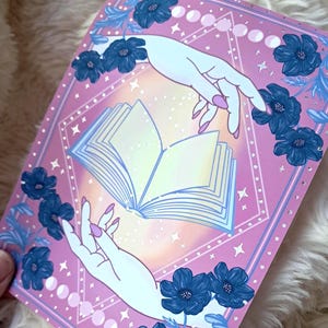 Puede incluir: Ilustraci&oacute;n rosa y azul con un libro abierto entre dos manos. La obra est&aacute; enmarcada con flores azules y fases lunares. Las manos tienen u&ntilde;as rosa claro. El fondo tiene un degradado de amarillo a rosa.