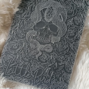 May include: A black and white graphic design of a skull with a snake wrapped around it. The design is surrounded by roses and the words "Little Bird" are visible at the bottom.