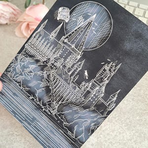 Op de afbeelding: Een zwart-witte illustratie van kasteel Zweinstein met een vliegende auto in de lucht. Het kasteel is omgeven door bomen en een meer. De illustratie is op een donkergrijze achtergrond.