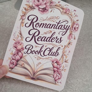 Może przedstawiać: Prostokątna karta z napisem "Romantasy Readers Book Club" w ozdobnej czcionce, otoczona różowymi ilustracjami kwiatowymi i grafiką otwartej książki. Karta ma białe tło.