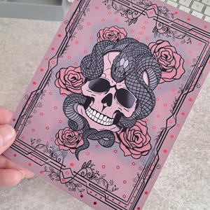 Może przedstawiać: Różowa i czarna ilustracja czaszki z wężem owiniętym wokół niej. Czaszka jest otoczona różami i ozdobną ramą z czerwonymi kropkami.