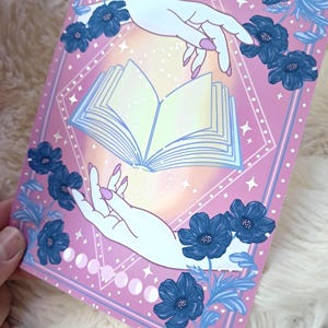 Puede incluir: Una impresi&oacute;n art&iacute;stica rosa con un libro abierto de p&aacute;ginas brillantes, sostenido por dos manos con u&ntilde;as rosas. Flores y hojas azules enmarcan la imagen, con fases lunares y estrellas. La obra tiene un tema m&iacute;stico.