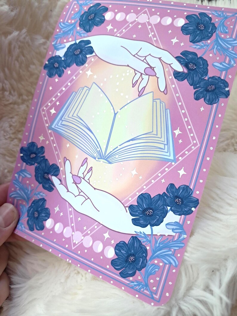 Puede incluir: Una impresi&oacute;n art&iacute;stica rosa con un libro abierto de p&aacute;ginas brillantes, sostenido por dos manos con u&ntilde;as rosas. Flores y hojas azules enmarcan la imagen, con fases lunares y estrellas. La obra tiene un tema m&iacute;stico.