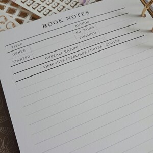 Pode incluir: Um livro branco com um padrão de grade preta e as palavras "BOOK NOTES" em texto preto. O livro tem seções para o título, o gênero, o início, o autor, o número de páginas, o final, a classificação geral e os pensamentos/sentimentos/notas/citações.